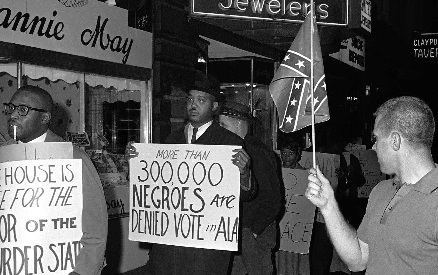 In this April 14, 1964, black-and-white file photo, a man holds a Confederate flag at right, as demonstrators, including one carrying a sign reading, “More than 300,000 Negroes are Denied Vote in Ala,” demonstrate in front of an Indianapolis hotel where then–Alabama Governor George Wallace was staying.