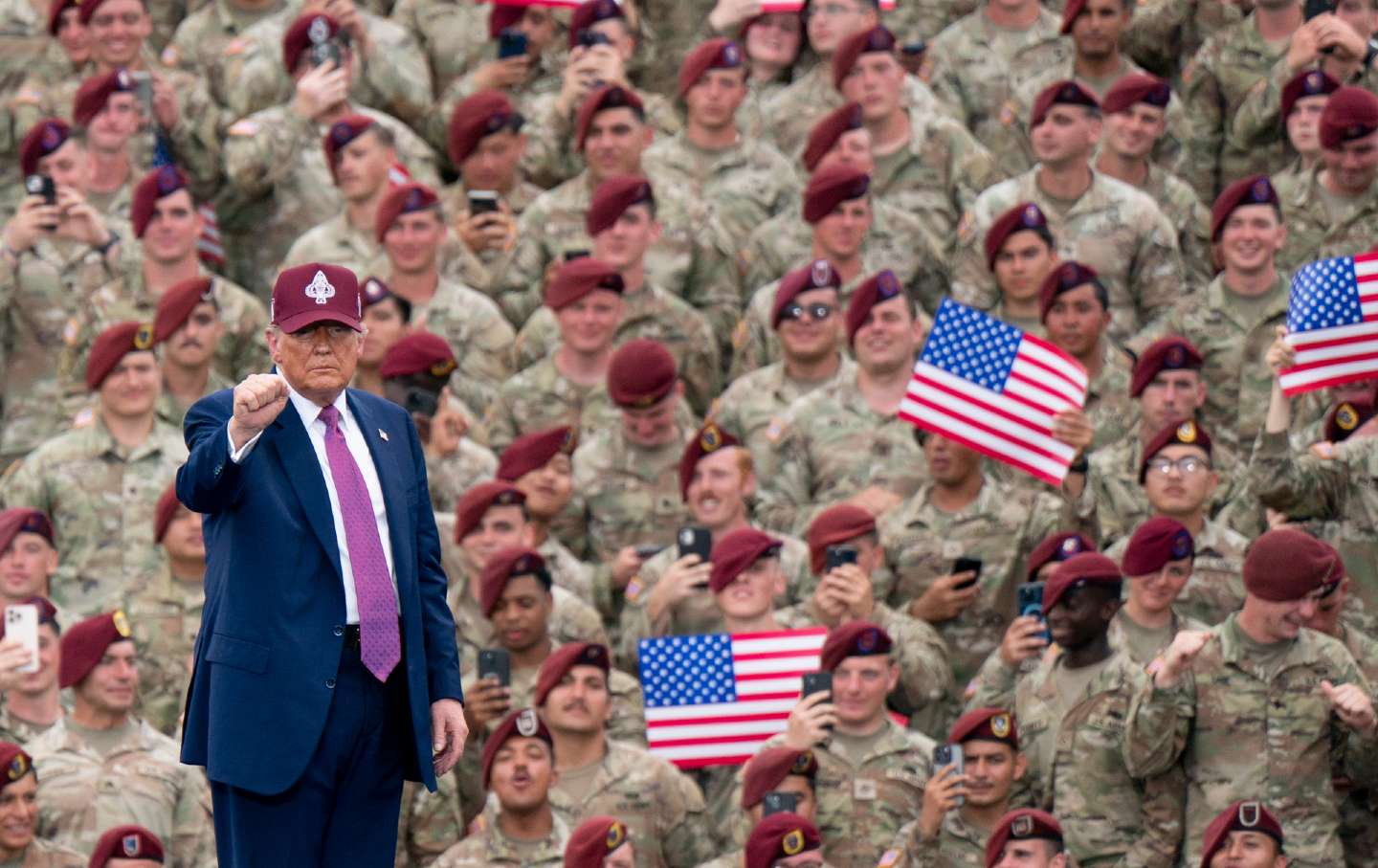 President Donald Trump wears a baseball cap with military insignia and holds up a fist. He faces away from a crowd of troops at Fort Bragg who wear camouflage-printed fatigues and maroon berets. Some troops hold up their phones or American flags.